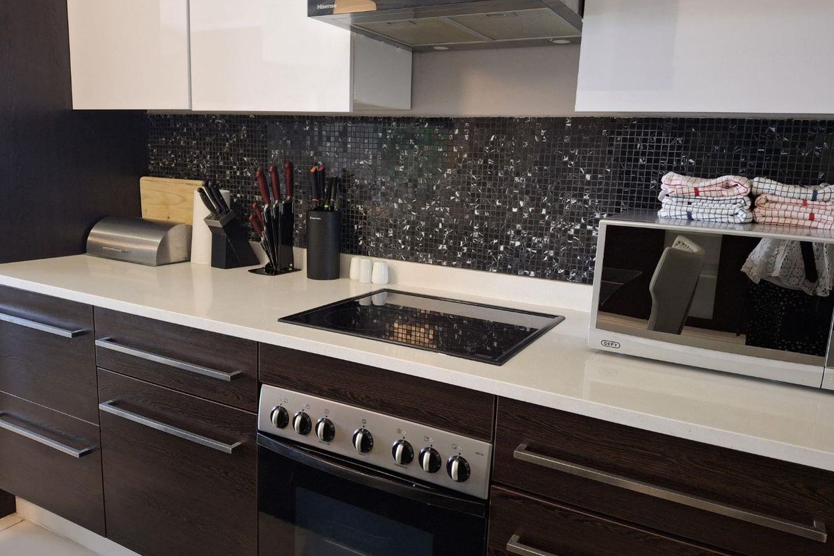 Modern kitchen featuring dark wood-grain cabinetry, white quartz countertops, a mosaic tile backsplash, and integrated stainless steel cooking appliances.