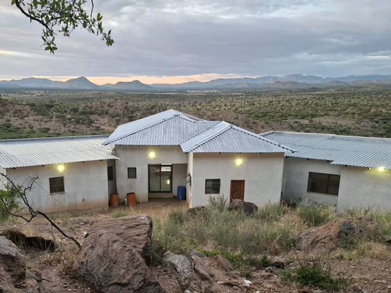 Rear view of a single-story house with a corrugated metal roof, plastered walls, and expansive mountain landscape views.