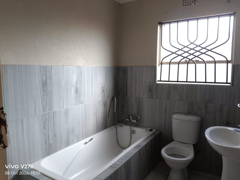 A functional bathroom featuring a bathtub, pedestal basin, and toilet, finished with wood-look wall tiles and fitted with security bars on the window.