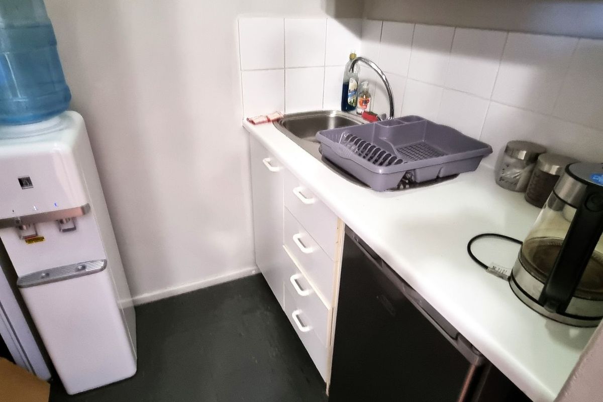 A compact kitchenette area featuring a built-in sink, tiled splashback, and under-counter cabinetry, suitable for office tea and coffee preparation.