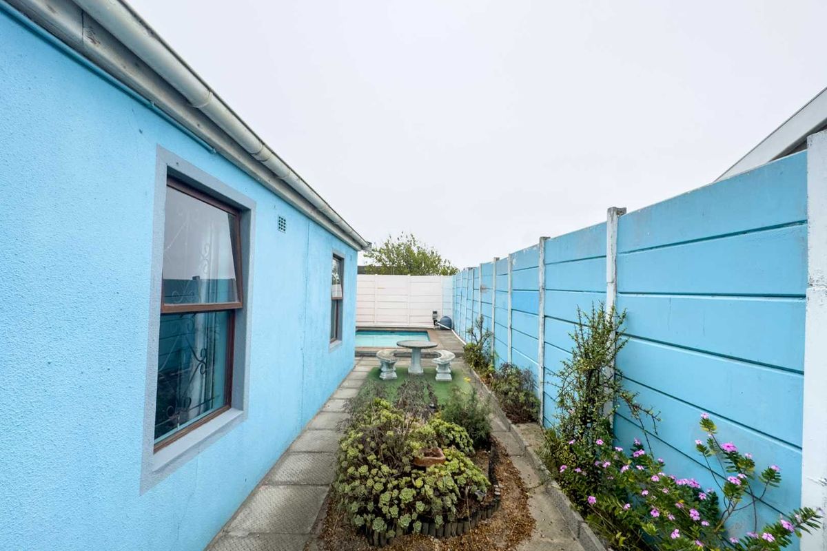 A narrow side garden area featuring a concrete pathway, perimeter boundary wall, and a glimpse of a swimming pool at the rear.