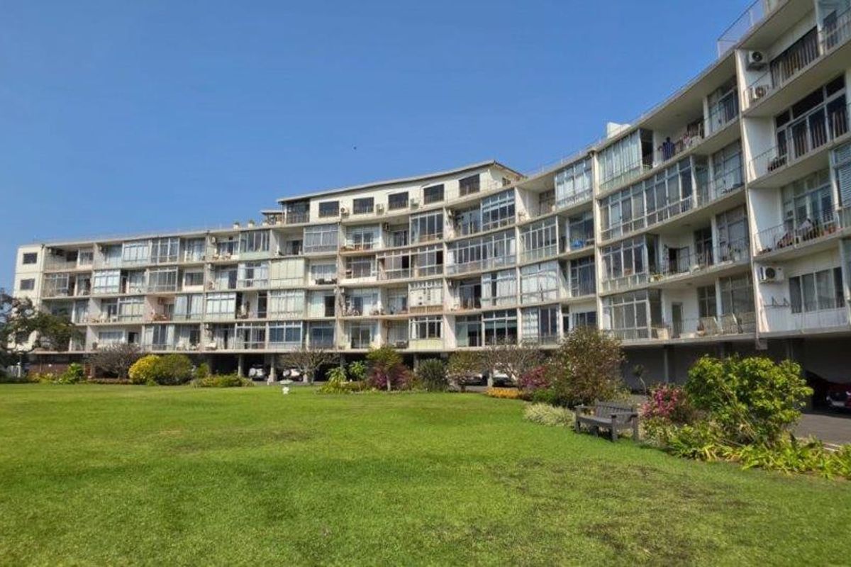 A view of the rear elevation of a multi-story apartment building featuring private balconies and ground-level covered parking overlooking a communal garden area.