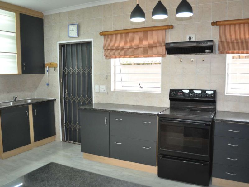 A functional kitchen featuring dark cabinetry, laminate countertops, a freestanding stove with extractor fan, and tiled splashbacks with a security gate entrance.