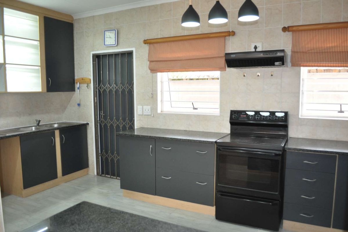 A functional kitchen featuring dark cabinetry, laminate countertops, a freestanding stove with extractor fan, and tiled splashbacks with a security gate entrance.