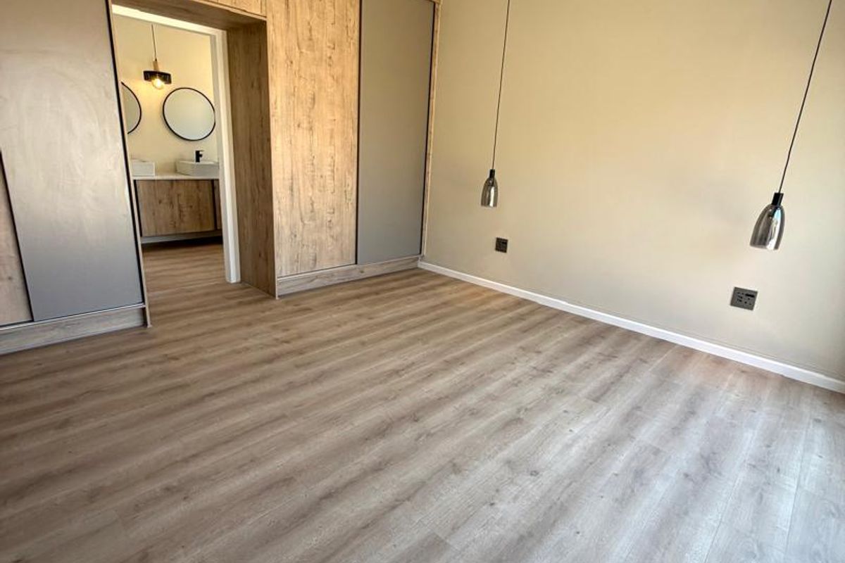 A modern, spacious bedroom featuring wood-look laminate flooring, built-in floor-to-ceiling cabinetry, contemporary pendant lighting, and an adjoining en-suite bathroom.
