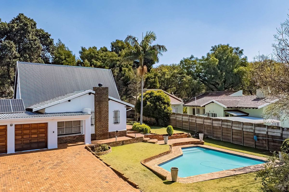 Aerial view of the rear property featuring a swimming pool, brick paved driveway, garage, and solar panels set within a landscaped garden.