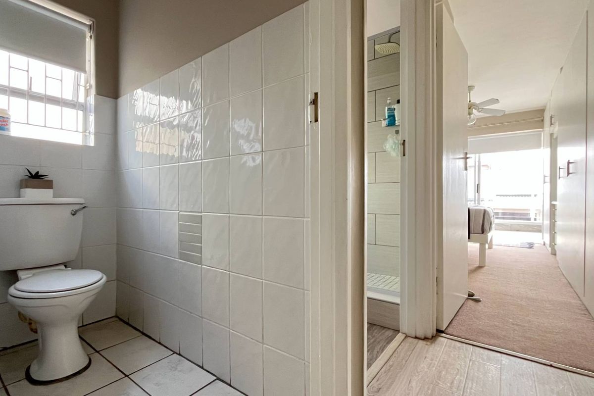 A functional bathroom featuring white wall tiles and floor tiles, with an adjacent view into a bedroom area with built-in storage.