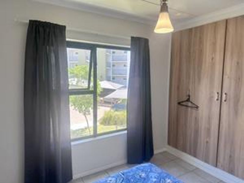 A bedroom featuring built-in wooden cupboards, tiled flooring, and an aluminium window frame with views of the complex grounds.