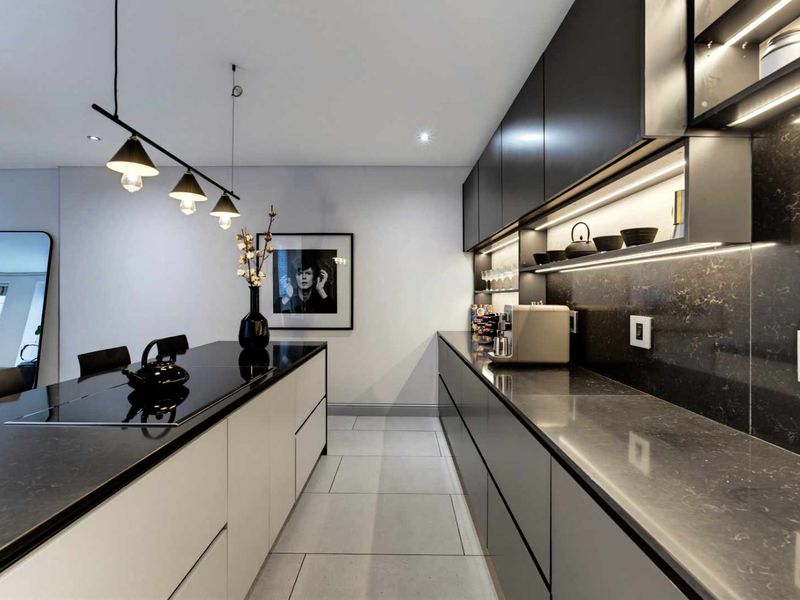 Modern kitchen featuring sleek dark cabinetry, stone countertops, integrated hob, and sophisticated under-cabinet lighting in a contemporary apartment setting.