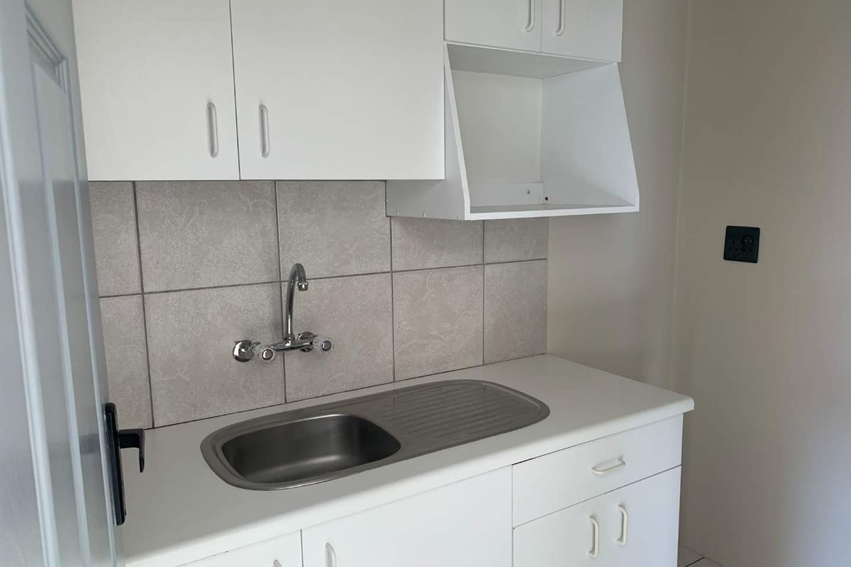 A compact kitchenette featuring white cabinetry, a stainless steel sink with wall-mounted faucet, and neutral tiled splashback.