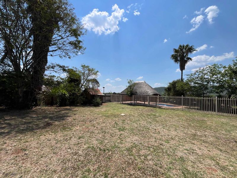 Spacious garden area featuring a swimming pool, a traditional thatched lapa, and established trees, enclosed by metal perimeter fencing.