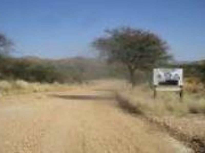 A view of a rural dirt road leading towards a landscape with trees and hills, featuring a roadside sign.