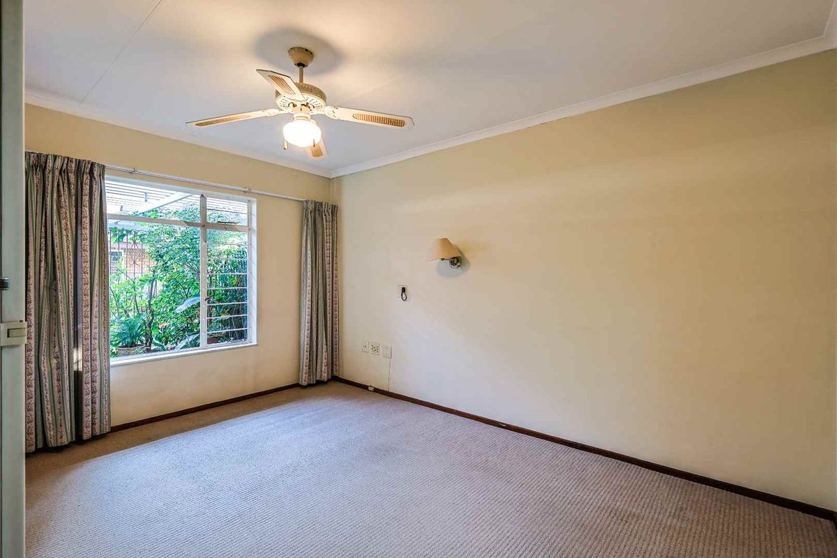 A spacious, neutral-toned bedroom featuring a ceiling fan, wall-mounted lighting, and a large window overlooking the garden.