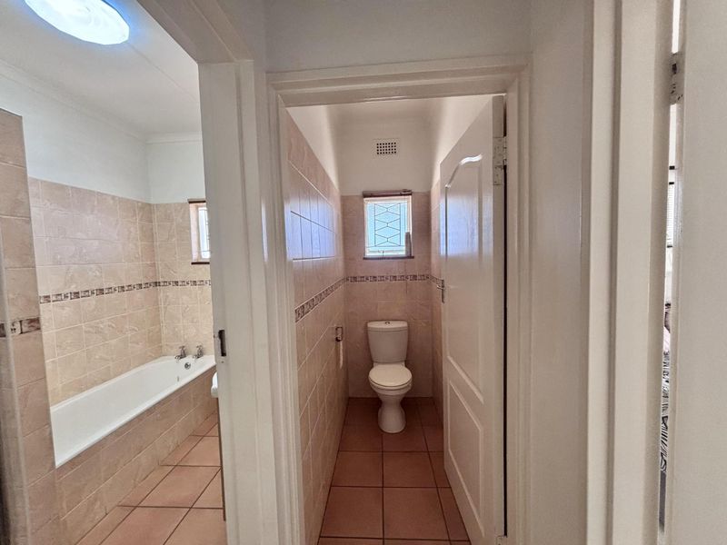 A functional bathroom featuring a built-in bathtub and a separate toilet area with neutral ceramic tiling throughout.