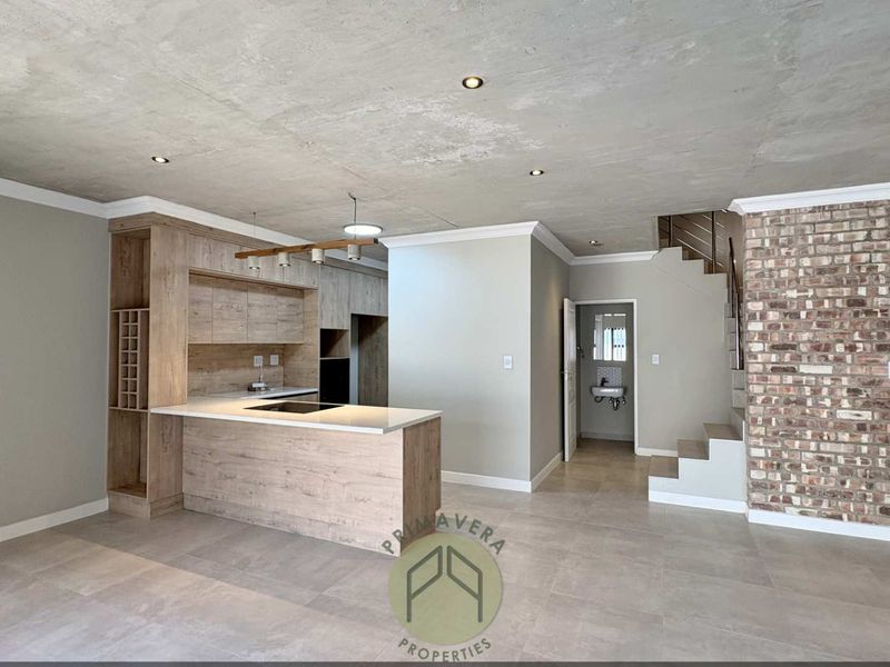 Modern open-plan living area featuring an exposed concrete ceiling, sleek kitchen cabinetry with a stone island, and a striking exposed brick feature wall.