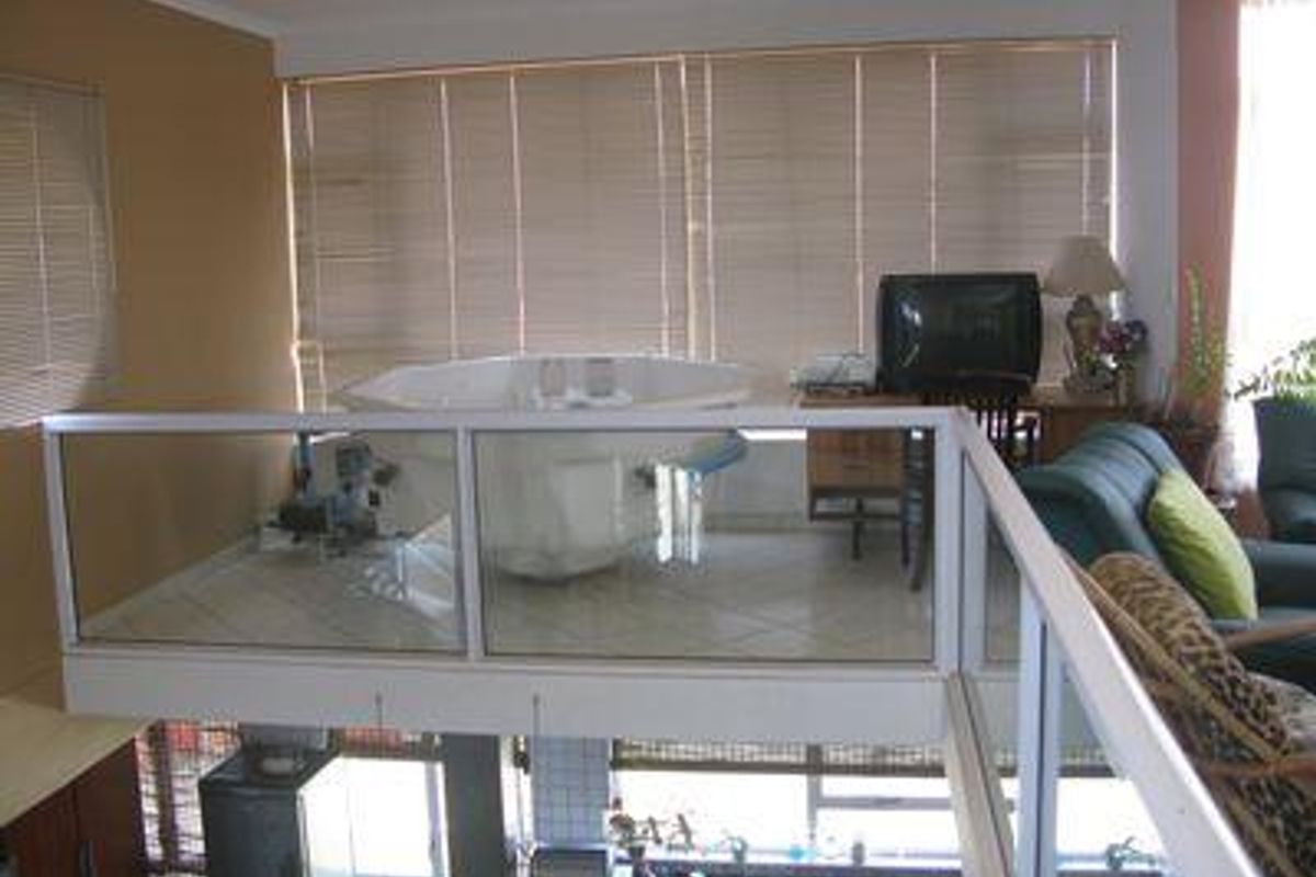 An elevated mezzanine bedroom area featuring a built-in bathtub, tiled flooring, and a glass balustrade overlooking the lower level.
