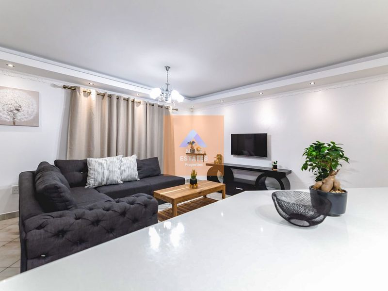 A well-maintained living room featuring recessed ceiling lighting, decorative cornice detailing, and neutral tiled flooring, creating a bright and comfortable living space.