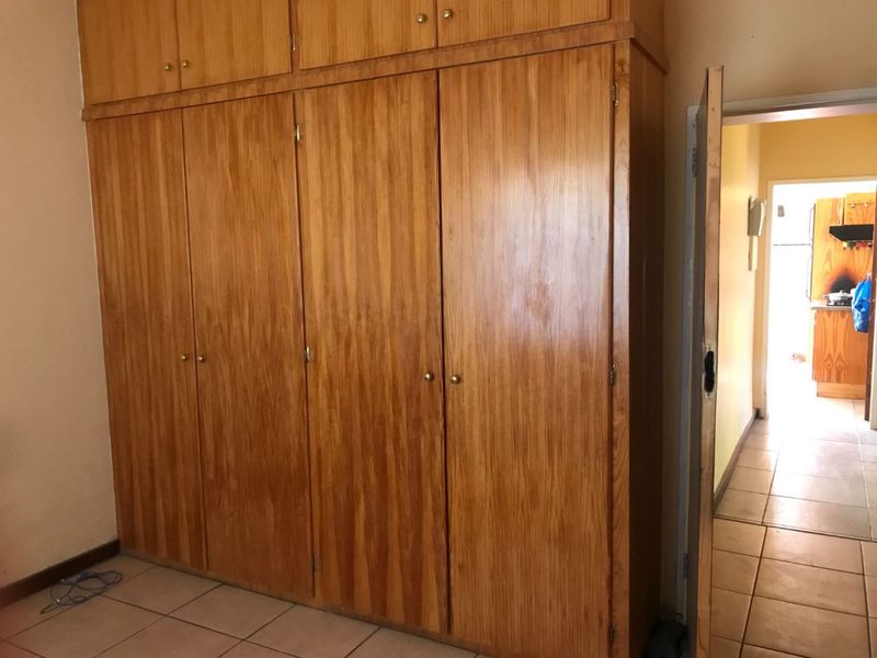 A bedroom featuring extensive floor-to-ceiling built-in wooden cupboards and tiled flooring, with a view through the doorway into the adjacent kitchen area.