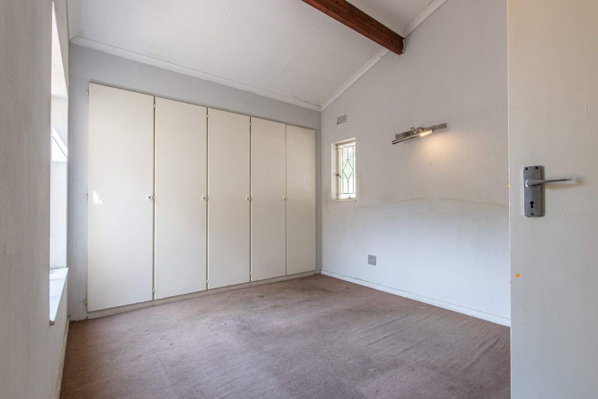 A vacant bedroom featuring built-in cupboards, a raked ceiling with exposed timber beam, and a small window with security bars.