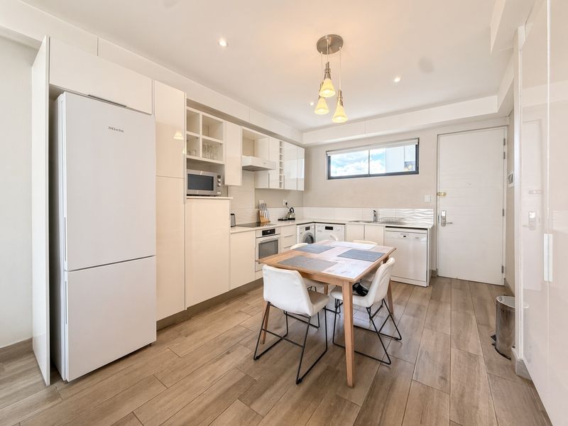 Modern open-plan kitchen and dining area featuring sleek white cabinetry, integrated appliances, tiled flooring, and contemporary lighting fixtures.
