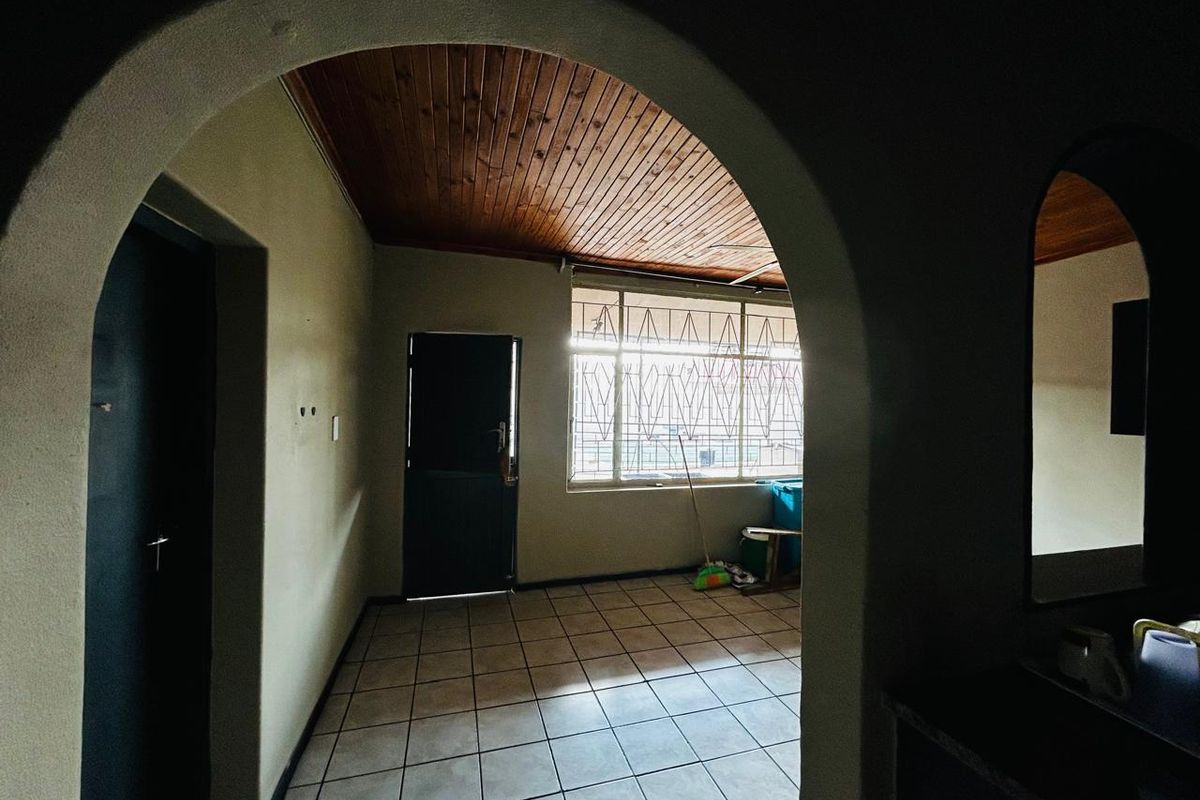 An interior view of a room featuring a distinctive arched doorway, tiled flooring, and a timber-paneled ceiling with security bars on the window.