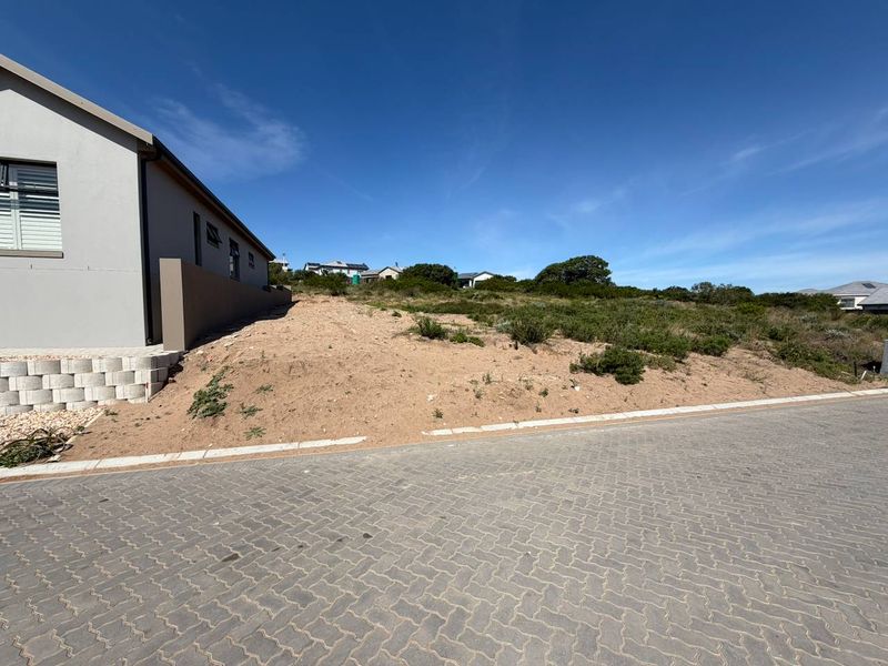 A view of a vacant residential plot featuring a paved street frontage and a sloped landscape with an adjacent retaining wall.