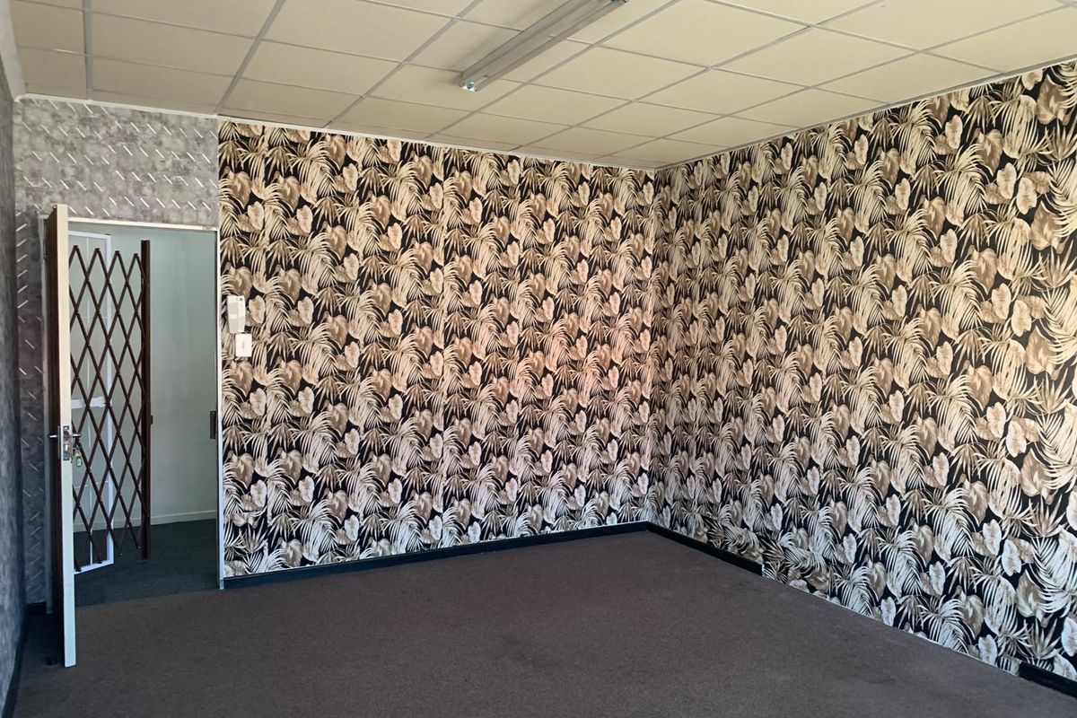 An empty office space featuring a suspended ceiling, patterned wallpaper, and carpet flooring, accessible via a security gate.