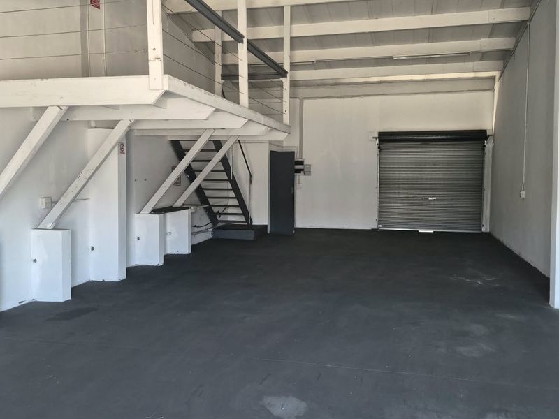Spacious industrial utility area featuring a mezzanine level, metal staircase, roller shutter door, and exposed timber rafters with durable concrete flooring.
