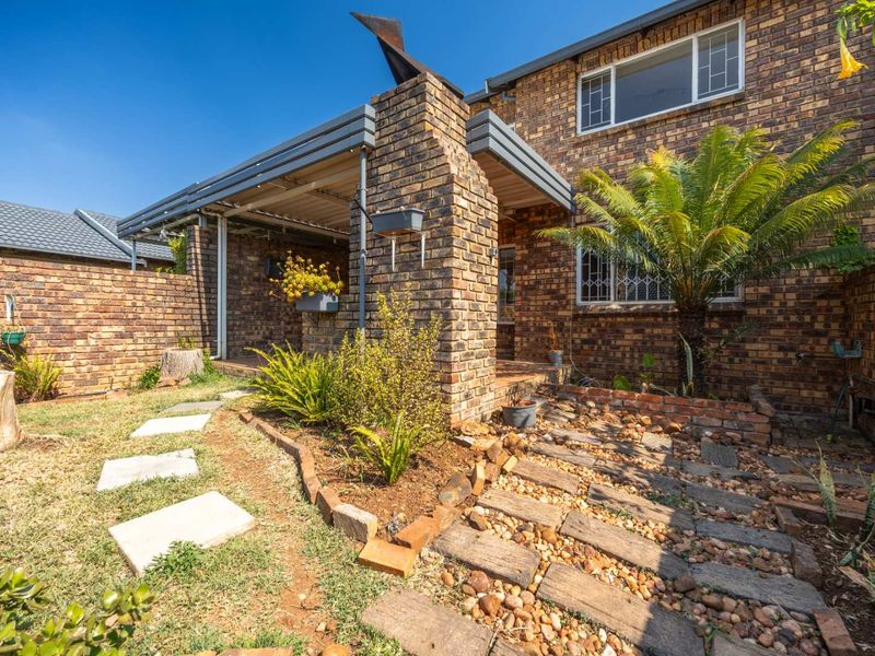 A rear exterior view of a face brick cluster home featuring a covered patio, paved garden pathways, and established landscaping.