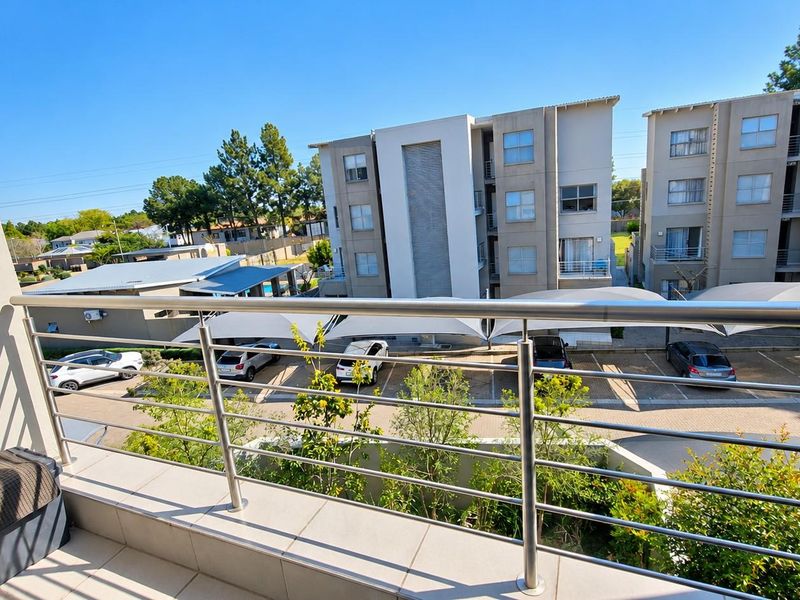 A clean, tiled balcony featuring a modern stainless steel balustrade, offering an elevated view over the apartment complex parking area and surrounding grounds.