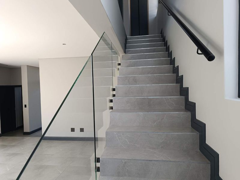 Modern staircase featuring grey stone-look porcelain tiles, a sleek frameless glass balustrade, and a minimalist black handrail in a bright, open hallway.