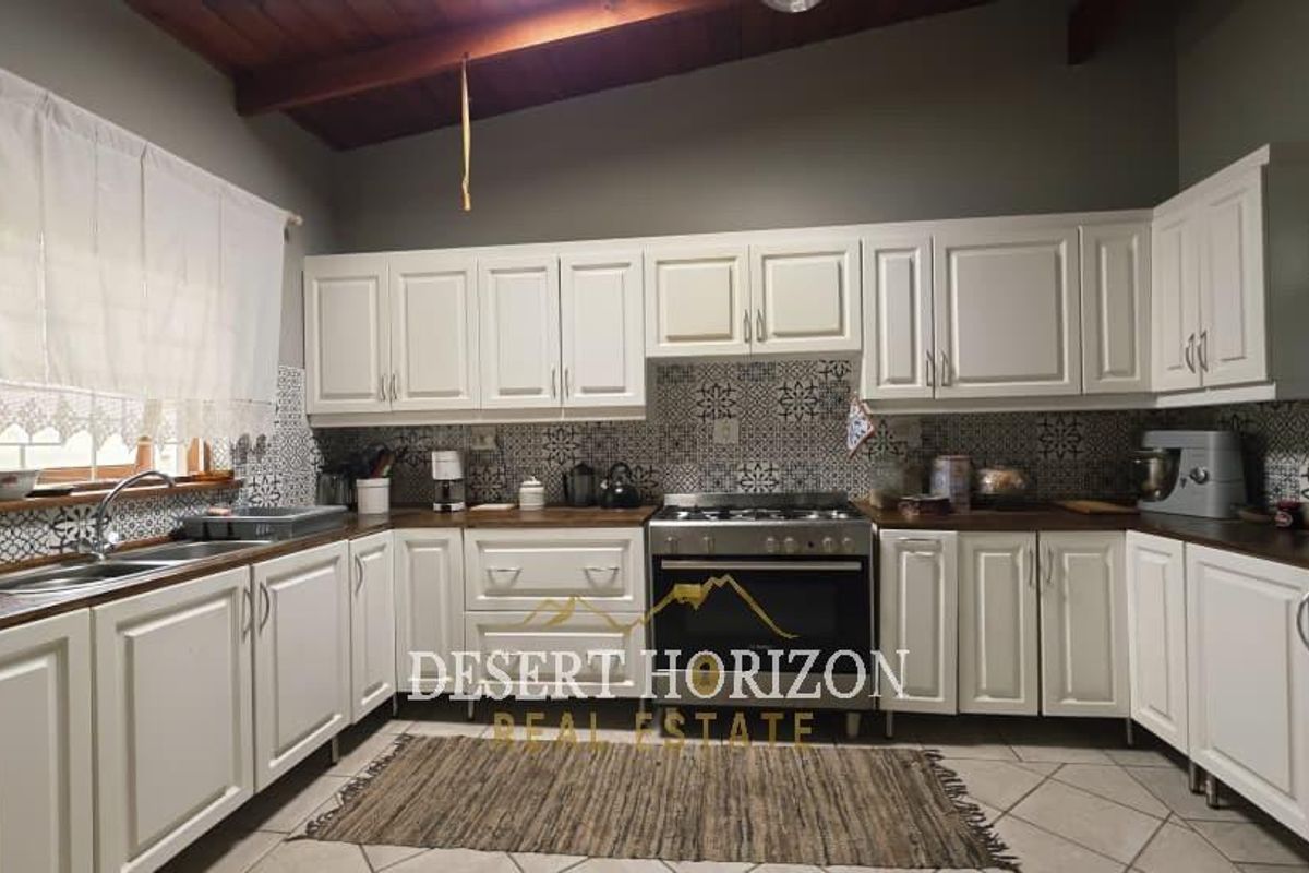 A well-maintained kitchen featuring white cabinetry, patterned tile backsplash, dark countertops, and a built-in gas stove with an exposed timber beam ceiling.