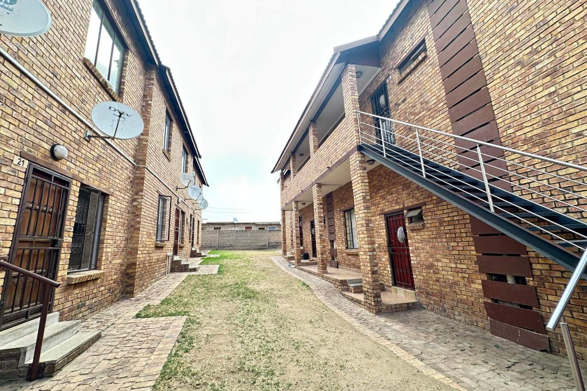 Exterior view of a face brick apartment complex featuring paved walkways, external staircases with metal railings, and communal garden space.