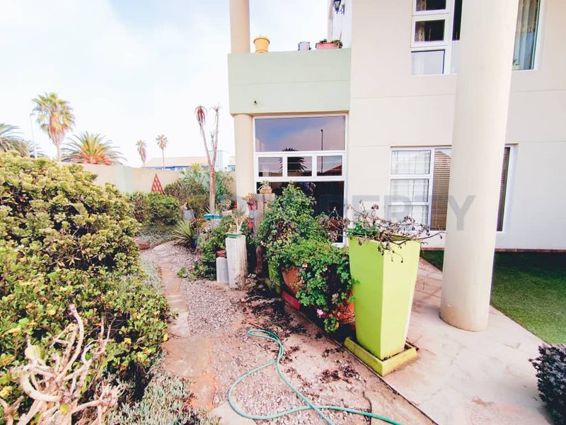 Rear exterior view of the apartment featuring a paved patio area, stone pathway, and established garden beds with structural columns.
