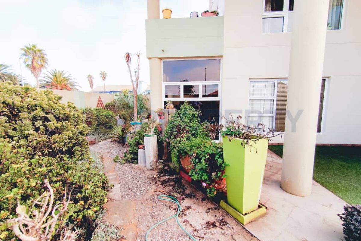 Rear exterior view of the apartment featuring a paved patio area, stone pathway, and established garden beds with structural columns.