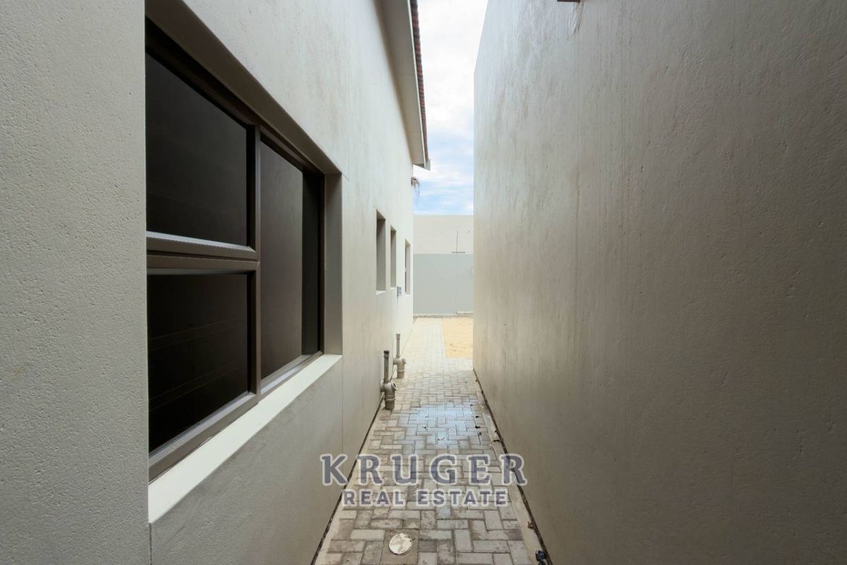 A narrow paved side passage between two exterior walls, featuring aluminium window frames and visible external plumbing infrastructure.