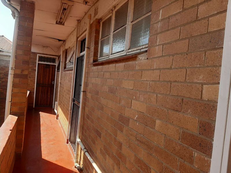 An exterior-facing apartment corridor featuring exposed brick walls, security gates, and timber doors, showing signs of significant wear and maintenance requirements.