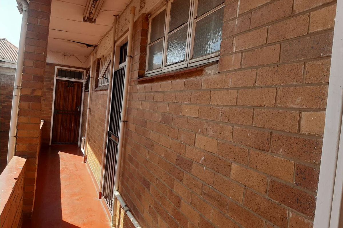 An exterior-facing apartment corridor featuring exposed brick walls, security gates, and timber doors, showing signs of significant wear and maintenance requirements.