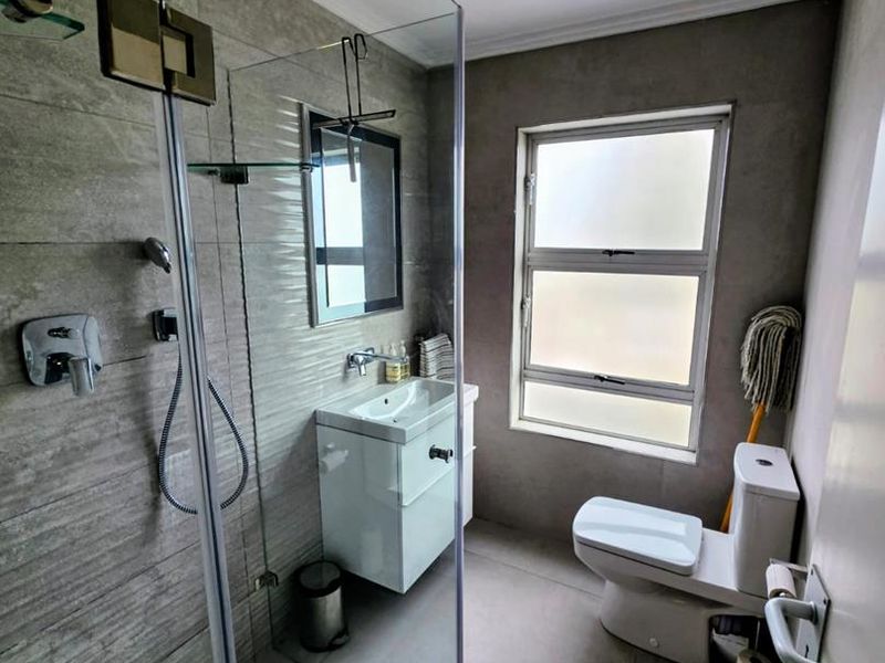 Modern bathroom featuring a glass-enclosed shower with rain shower head, vanity basin, and toilet, finished with contemporary grey wall and floor tiles.