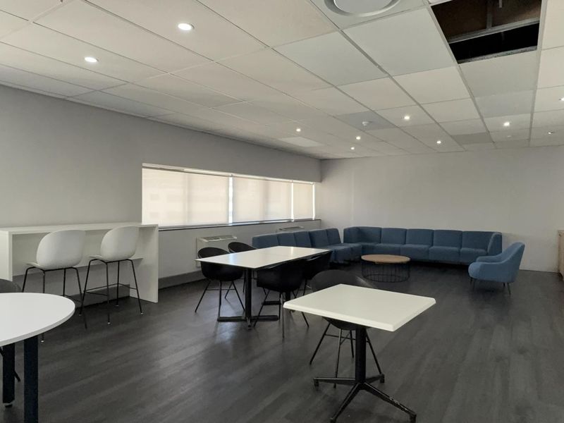 A spacious office break-out area featuring dark laminate flooring, a suspended ceiling with recessed lighting, and large windows with fitted blinds.