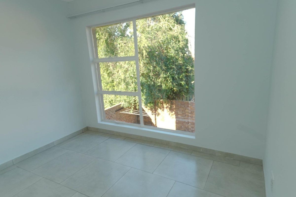 A bright, empty bedroom featuring large windows with a garden view and neutral tiled flooring.