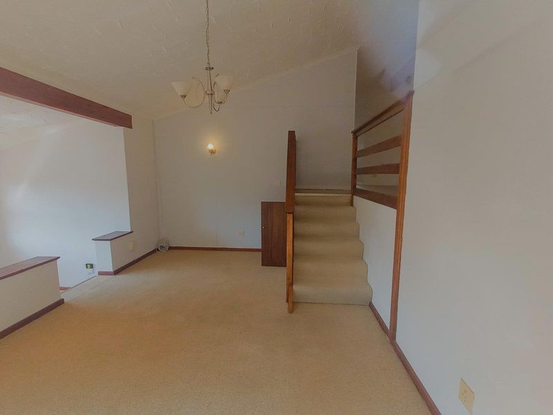 An empty living area featuring a staircase with a wooden balustrade, carpeted flooring, and exposed ceiling beams.