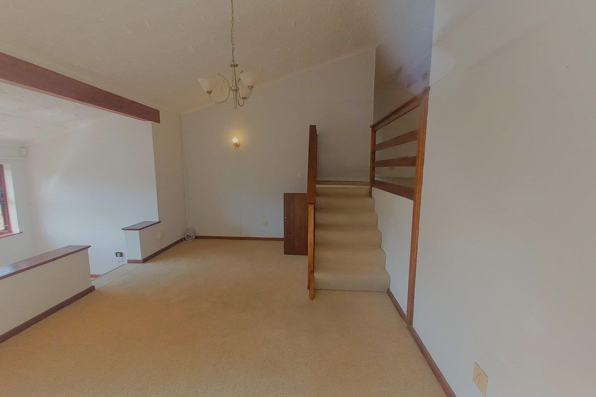 An empty living area featuring a staircase with a wooden balustrade, carpeted flooring, and exposed ceiling beams.