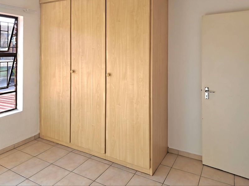 A clean, empty bedroom featuring light wood-finish built-in wardrobes, neutral tiled flooring, and a window with security bars.