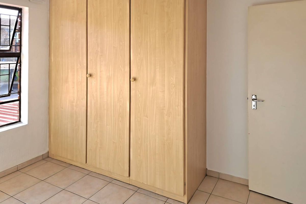 A clean, empty bedroom featuring light wood-finish built-in wardrobes, neutral tiled flooring, and a window with security bars.