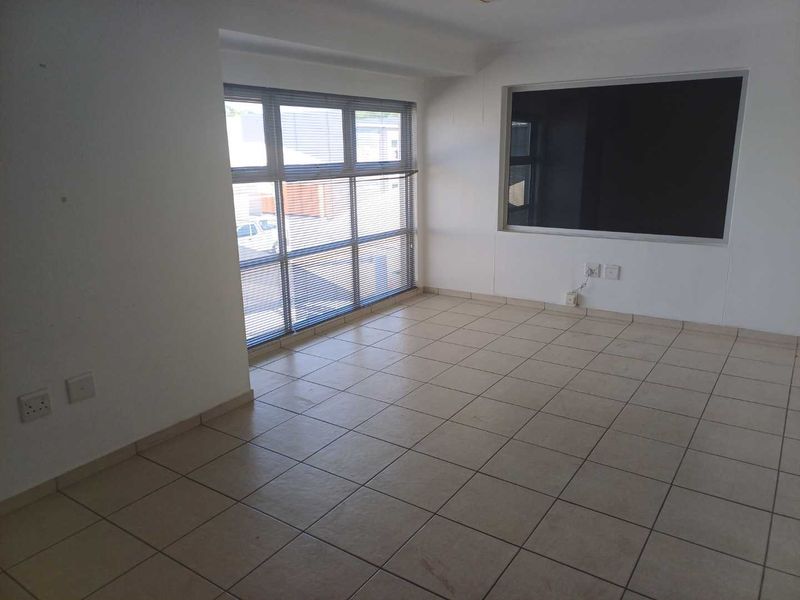 A bright, empty office space featuring neutral tiled flooring, large windows with venetian blinds, and a built-in display panel.