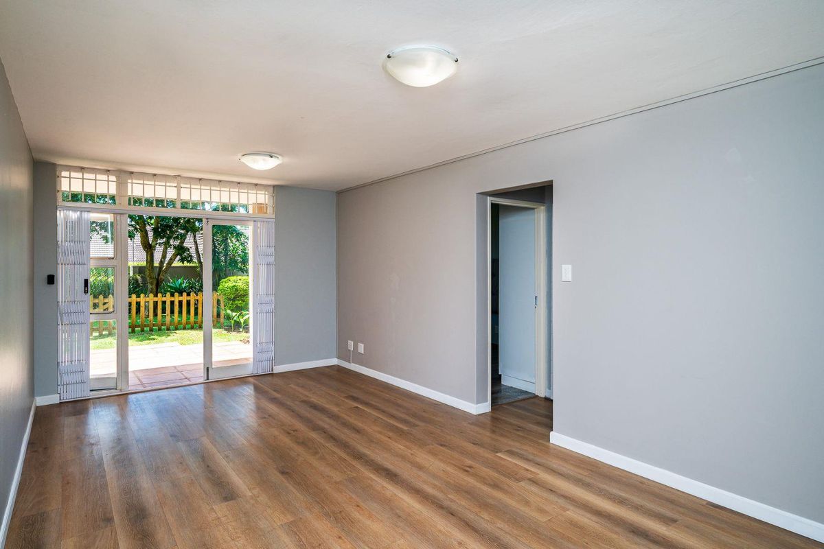A bright, empty living room featuring modern laminate flooring, neutral grey walls, and a sliding glass door providing direct access to the garden.