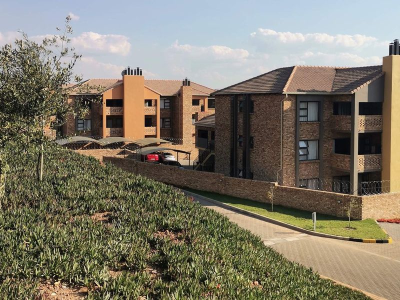 Exterior view of a multi-story apartment complex featuring brick facades, tiled roofing, and secure perimeter fencing with a paved driveway and carport area.