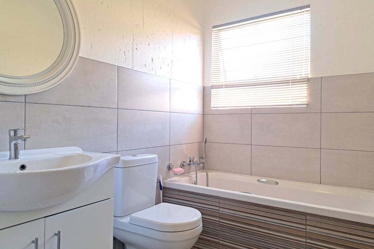 A functional bathroom featuring a white vanity, toilet, and a bathtub with decorative tiled side paneling and a window with blinds.