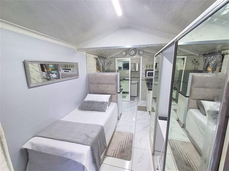 A compact bedroom featuring tiled flooring, a vaulted ceiling, and a large mirrored sliding wardrobe, with an adjoining view into a small kitchenette area.